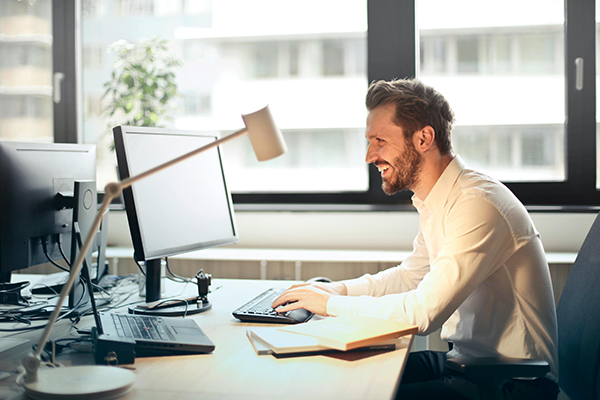 A man in a white shirt sits at a desk with multiple computer monitors, typing and smiling. A desk lamp, notebook, and pen are also visible.